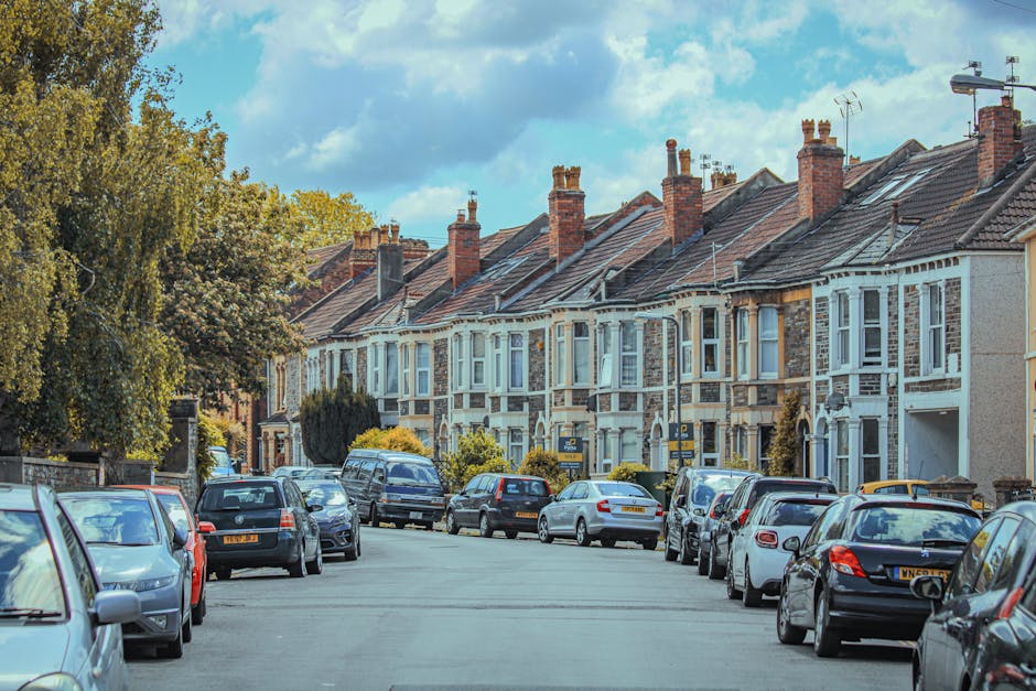 Photograph of a residential street in Neasden with a row of terraced houses featuring brick and stucco facades, bay windows, and tiled roofs with multiple chimneys. Several cars are parked along both sides of the street, including small hatchbacks, sedans, and a van. In the foreground, a man from Man with Van Neasden is seen loading or unloading a large cardboard box with packing materials, using a dolly to move it towards the van parked on the pavement. The area is well-lit with natural daylight, and there are trees with green and yellow foliage on the left side, indicating an autumn setting. The scene captures the home relocation process, focusing on furniture transport and packing and moving activities typical of house removals, with the clear environment and accessible layout suitable for managing moving logistics.