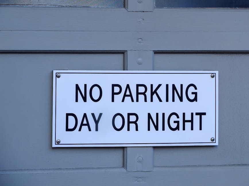 A circular traffic sign with a red border and a blue interior featuring a red diagonal line indicating no parking is mounted on a metal pole, casting a shadow on a textured brick wall composed of light and dark stones with a horizontal red brick stripe near the bottom. The sign is positioned near a property entrance or street scene involved in house removals or furniture transport, with natural lighting highlighting the sign and wall surface. This image relates to the planning needed for home relocation and moving logistics, as it emphasizes parking restrictions that can impact the loading process for a professional removals service such as Man with Van Neasden, especially on a moving day to avoid parking suspensions.
