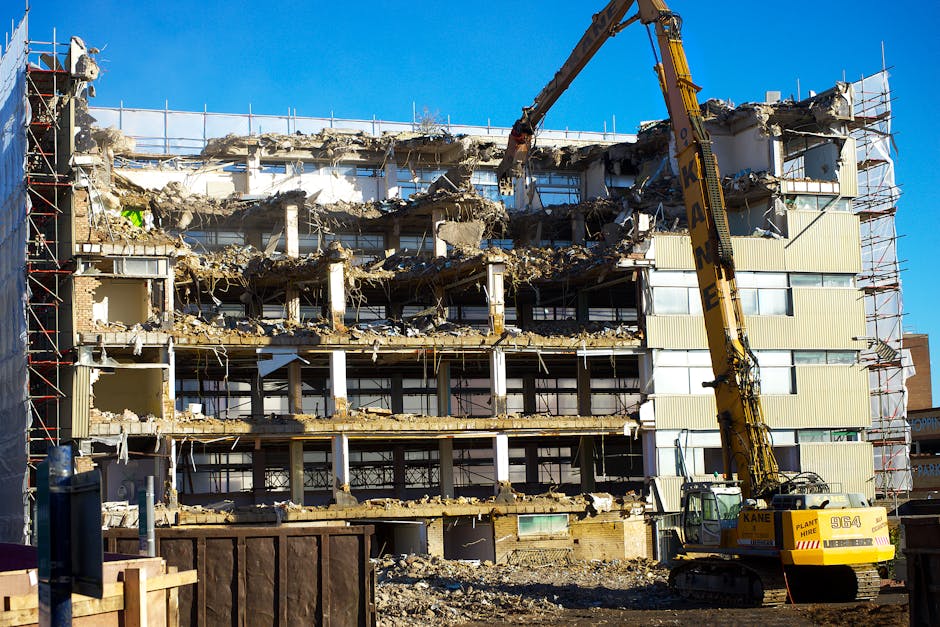 A multi-storey building is undergoing demolition in an urban area, with sections of exterior walls and floors partially torn down, exposing the building’s interior structure. A large yellow demolition crane with an extended arm and claw attachment is actively dismantling the upper floors, with debris spilling onto the lower levels. Scaffolding surrounds the building’s facade on the right side, reinforcing the ongoing construction work. The ground around the site is cluttered with rubble, broken concrete, and discarded materials, while a temporary wooden barrier is positioned in the foreground. Bright daylight illuminates the scene against a clear blue sky, highlighting the raw and chaotic nature of the demolition process, which is part of a property development or refurbishment project. Occasionally, [COMPANY_NAME] services are involved in such building works, supporting safe and efficient demolition and material removal, aligned with house removals and construction logistics.
