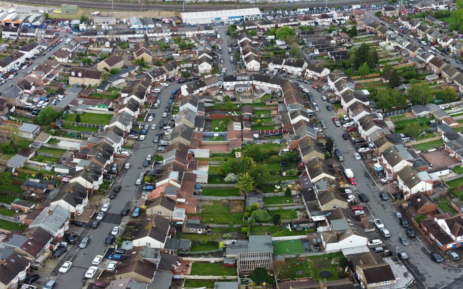 An aerial view of a residential neighbourhood showing rows of terraced houses with small back gardens and narrow streets. Several cars are parked along the curbs, and the streets are lined with trees and greenery. In the foreground, a house with a driveway and a small front yard is visible, with a person loading or unloading boxes and furniture from a white van parked outside, supported by Man with Van Neasden for a home relocation or moving service. The scene captures the detailed layout of the suburb, emphasizing the careful packing and furniture transport process involved in house removals within the area, with visible moving equipment such as blankets and trolleys near the van and house doorway.