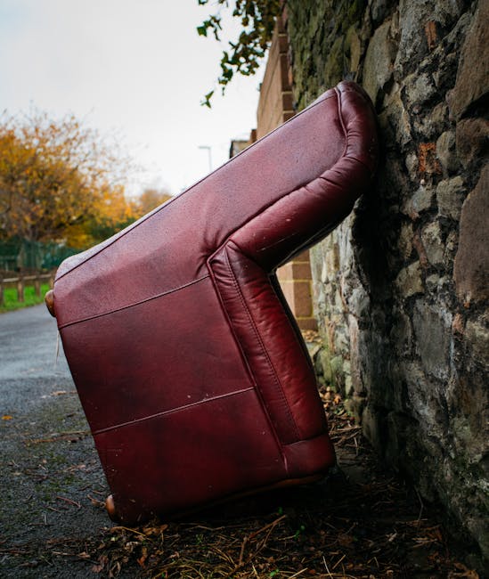 A red leather armchair has been positioned upside down on a paved outdoor surface, leaning against a textured brick and stone wall. The chair's legs are pointing upwards, while the seat and backrest rest at an angle on the ground. In the background, there are trees with autumn foliage, a green fence, and a paved pathway, suggesting an outdoor area near a residential property. The scene appears to depict a furniture removal process, possibly involving loading or unloading in preparation for a home relocation. The lighting is natural, indicating daytime, and the surrounding environment is quiet and residential, consistent with typical house removals and furniture transport situations. The image aligns with the services offered by Man with Van Neasden, focusing on furniture collection and home moving logistics.