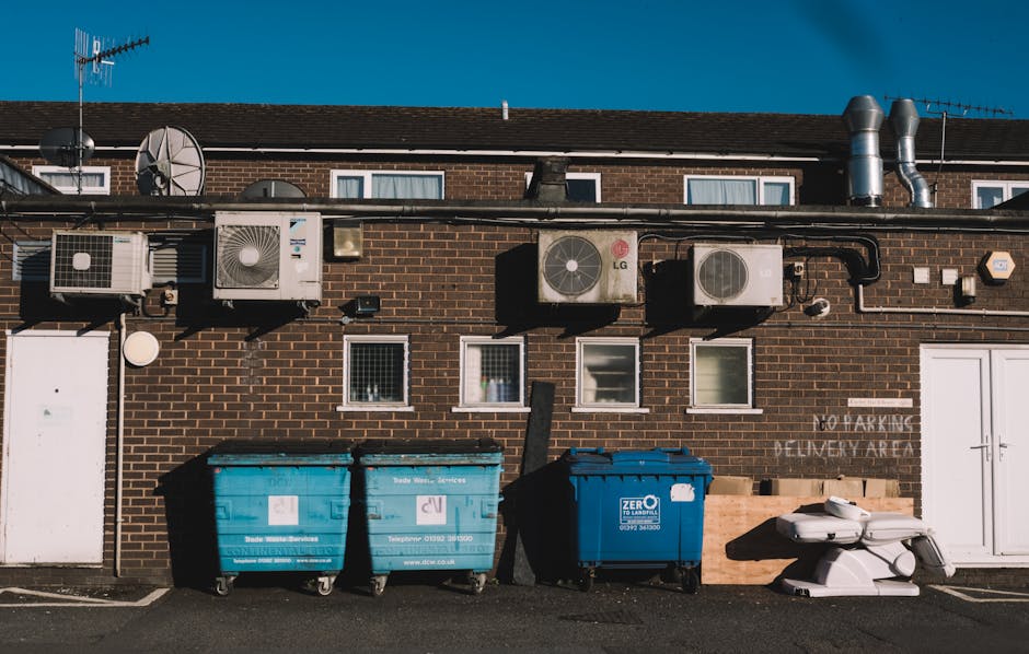 A white garbage collection truck with the number 610 on its side is parked on a street in Neasden, near a row of brick residential buildings with multiple windows and small balconies. The truck is loaded with various waste materials, including cardboard boxes, plastic bags, and an orange container, which are stacked on top of each other on the truck's rear. The truck's large rear compartment is tilted slightly backward, indicating an active loading or unloading process. In front of the truck, on the pavement, there are several black trash bags and loose waste items, suggesting ongoing collection of bulky waste or household rubbish. The scene is illuminated by natural daylight, with trees casting shadows over the buildings and the vehicle, and the environment appears calm and organized, reflecting typical waste collection activities in an urban residential area. Occasionally, Man with Van Neasden's services facilitate such home relocation and waste removal processes, supporting efficient moving logistics.