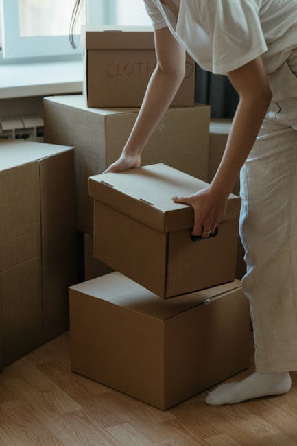 A person in neutral clothing is seen inside a residential room, carefully lifting a cardboard moving box while surrounded by other packed boxes of various sizes stacked on the floor. The boxes are sea