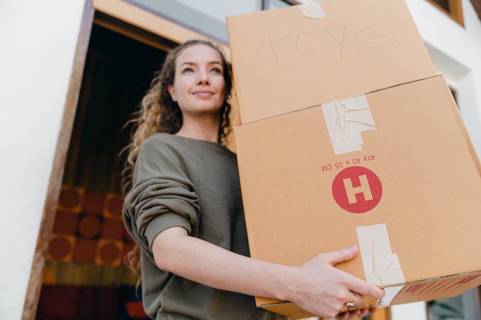 A person in neutral clothing is seen inside a residential room, carefully lifting a cardboard moving box while surrounded by other packed boxes of various sizes stacked on the floor. The boxes are sea