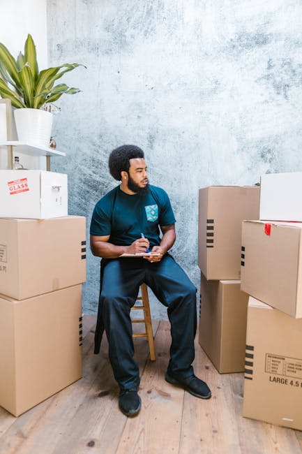 A man with dark curly hair and a beard, wearing a blue uniform with a logo on the chest, is seated on a wooden stool inside a room with a light textured wall. He is surrounded by stacked cardboard boxes, some sealed with tape, indicating packing for a home relocation or furniture transport. To his left, a white shelf holds a potted plant with broad green leaves and a white box labeled 'GLASS'. The boxes vary in size, with some on the floor and others on a small table or stacked against the wall, ready for loading or unloading. The wooden floor and neutral wall create a bright environment, suitable for describing a professional packing and moving process. These visual details support the services of Man with Van Neasden, highlighting the packing and logistics phase of house removals and furniture transport.