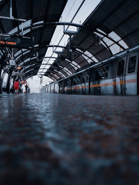 The image shows a modern train station platform with a curved, transparent roof structure supported by metal beams, allowing natural light to illuminate the area. A silver train with an orange stripe along its side is parked along the platform, with several windows visible. On the platform, there are a few passengers, some standing and others walking, carrying luggage or backpacks. To the left, there is a sign indicating the destination and platform information, with a digital display showing details. The textured surface of the platform floor, possibly made of concrete or similar material, is in the foreground and appears slightly wet, adding to the realistic, everyday scene of a typical London commuter station. This setting can relate to home relocation plans or moving logistics involving transportation and travel, and is related to [COMPANY_NAME]'s removals and moving services in the Neasden area.