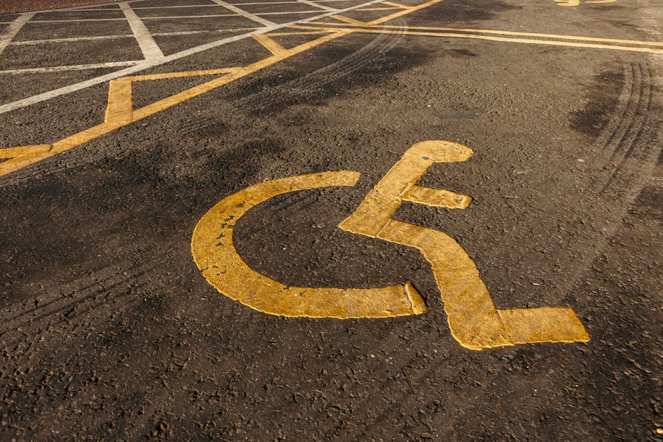 Close-up view of a pedestrian parking space designated for disabled persons, painted with a yellow wheelchair symbol on a dark asphalt surface. The parking bay is marked with yellow lines next to the symbol, indicating accessible parking area in a public or commercial parking lot. The surface shows minor wear and tyre marks, suggesting regular use. The background includes additional parking markings and spaces, with part of a moving van or vehicle potentially visible nearby, hinting at a home relocation or furniture transport scene. The image emphasizes the importance of accessible parking during house removals or moving services, which [COMPANY_NAME] provides in the Neasden area, assisting clients in efficient and accessible home moves. Proper access and parking considerations are crucial for smooth loading and unloading processes, especially near residential properties like those near Neasden Temple.