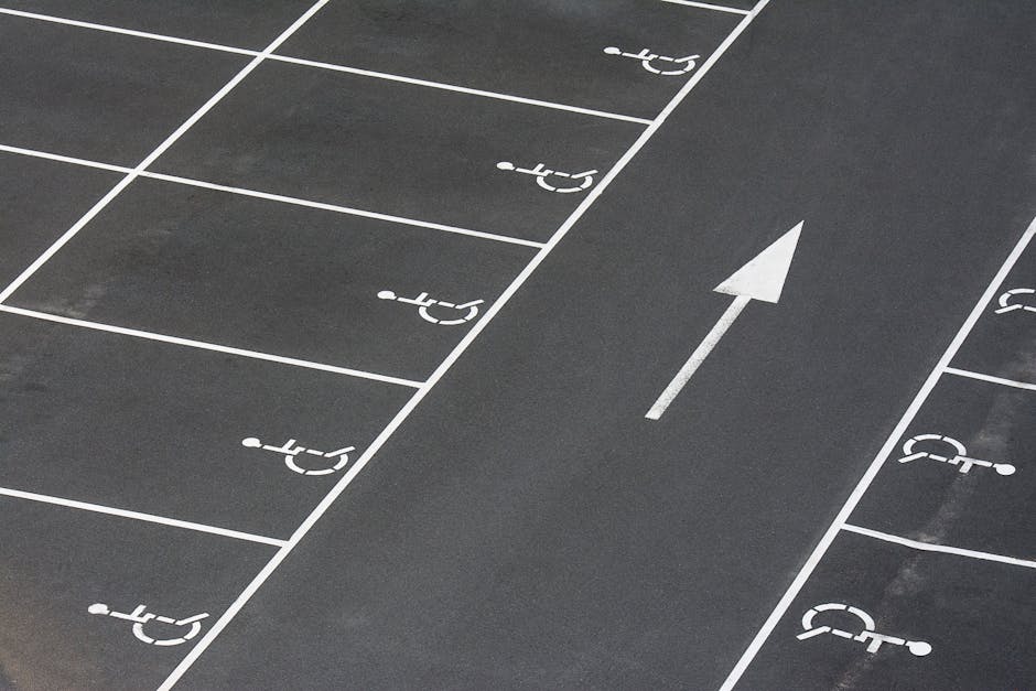 A section of a paved parking lot showing designated accessible parking spaces marked with white wheelchair symbols on the black asphalt surface. An arrow painted on the ground indicates the direction for vehicle movement. The parking spaces are separated by white lines, with some spaces located near the entrance of a property, likely used for moving trucks or service vehicles involved in home relocation or furniture transport. In the background, part of a loading or unloading area is visible, with no vehicles or objects present. The scene is well-lit, with clear markings supporting efficient parking and loading operations, relevant for professionals providing removals services such as those from Man with Van Neasden, as referenced on the specified webpage.