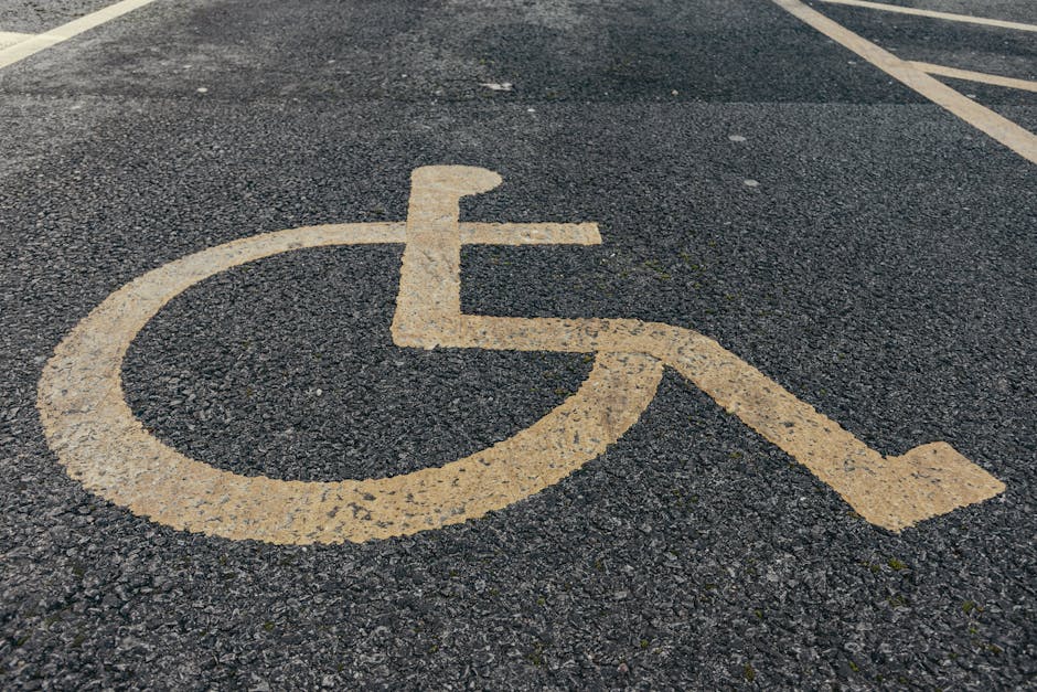 Close-up view of a pedestrian parking space designated for disabled persons, painted with a yellow wheelchair symbol on a dark asphalt surface. The parking bay is marked with yellow lines next to the symbol, indicating accessible parking area in a public or commercial parking lot. The surface shows minor wear and tyre marks, suggesting regular use. The background includes additional parking markings and spaces, with part of a moving van or vehicle potentially visible nearby, hinting at a home relocation or furniture transport scene. The image emphasizes the importance of accessible parking during house removals or moving services, which [COMPANY_NAME] provides in the Neasden area, assisting clients in efficient and accessible home moves. Proper access and parking considerations are crucial for smooth loading and unloading processes, especially near residential properties like those near Neasden Temple.