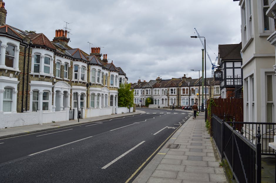 A quiet residential street in Neasden with a row of Victorian-style terraced houses featuring bay windows, brick exteriors, and tiled roofs. The street has a wide, paved sidewalk on the right side, with a black metal fence and a small garden area in front of one house. Overcast grey clouds cover the sky, creating soft, diffuse lighting. Several chimneys are visible on the rooftops, and a street lamp is positioned near the curb. In the distance, a blue crane is seen, indicating ongoing construction. The street is empty of moving vehicles or pedestrians, aside from a single person walking away from the camera. The scene captures a typical urban environment suitable for home relocation and furniture transport activities, consistent with the services provided by Man with Van Neasden for house removals and packing and moving processes.