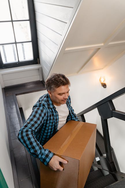 A young boy with curly red hair, dressed in a yellow plaid shirt over a white T-shirt, stands in a hallway holding a framed photograph in one hand and a potted plant with broad green leaves in the other. The hallway has plain white walls and is lit with natural lighting. Behind him, a staircase with dark wooden steps and gray railings is partially visible, indicating movement or transition within a home environment. The scene suggests preparations for a home relocation, with the boy possibly assisting in packing or moving household items. Nearby, there are cardboard boxes and packing materials, supporting the context of furniture transport and packing and moving activities, which are typical in house removals and relocation services offered by companies such as Man with Van Neasden. The overall setting reflects an indoor space where items are being organized or loaded for transport, illustrating aspects of the moving process within a residential property.