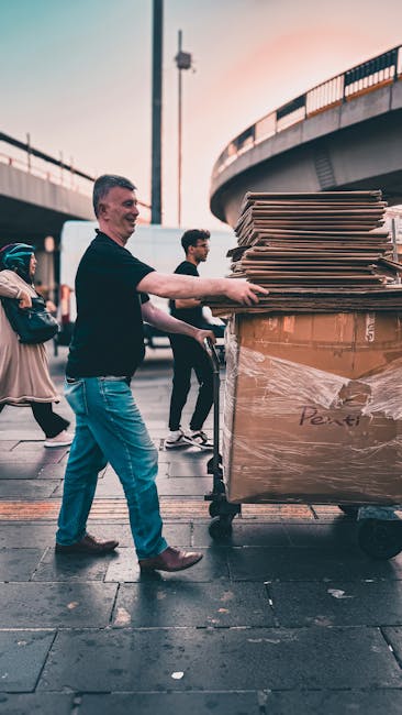 A man in a black shirt and blue jeans pushes a large, fully packed moving trolley loaded with flattened cardboard boxes and plastic-wrapped items, on a paved sidewalk under a bridge or elevated roadway. Several pedestrians are visible in the background, walking nearby. The scene is outdoors with natural lighting, capturing a moment during a home relocation or furniture transport process. The image reflects packing and loading activities typical of house removals, with a focus on efficient moving logistics as handled by Man with Van Neasden, a professional moving service. The surrounding environment includes urban infrastructure such as the underside of a bridge with metal railings and support structures, emphasizing the street-loading context of the moving operation.
