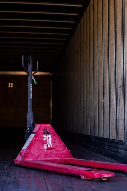 A red pallet truck or hand pallet jack positioned inside a storage or loading area, with the handle extended upward and the forks resting on the dark wooden floor. The surrounding environment includes wooden paneling on the right side wall and a ceiling with dark horizontal beams. The pallet truck is used for moving large or heavy items during home relocation or furniture transport, which may be part of a packing and moving process. This equipment is situated near a doorway or entry point for loading and unloading items for house removals, with natural light illuminating part of the space. The scene is neutral and professional, reflecting typical logistics and transport procedures carried out by Man with Van Neasden, supporting efficient load handling during street-loading operations in compliance with local regulations on moving in Neasden.