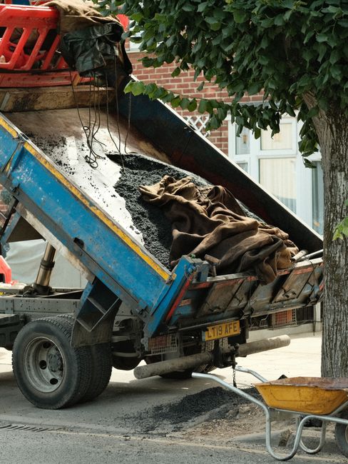 A man in a black shirt and blue jeans pushes a large, fully packed moving trolley loaded with flattened cardboard boxes and plastic-wrapped items, on a paved sidewalk under a bridge or elevated roadway. Several pedestrians are visible in the background, walking nearby. The scene is outdoors with natural lighting, capturing a moment during a home relocation or furniture transport process. The image reflects packing and loading activities typical of house removals, with a focus on efficient moving logistics as handled by Man with Van Neasden, a professional moving service. The surrounding environment includes urban infrastructure such as the underside of a bridge with metal railings and support structures, emphasizing the street-loading context of the moving operation.