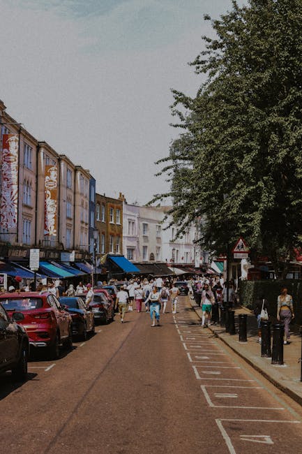 A busy street scene showing a row of multi-storey buildings with colorful facades, some featuring signage and awnings, lining a narrow road with parked cars on one side and space for moving vehicles o