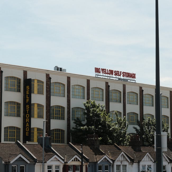 A multi-storey commercial building with a white facade and large arched windows framed in yellow on each floor, situated behind a row of small residential houses with pitched roofs. The commercial building has prominent signage reading 'SELF STORAGE' vertically along the side and 'BIG YELLOW SELF STORAGE' on a red sign at the top. There are trees partially obscuring the lower part of the building, and a lamppost is visible on the right side of the image. The scene appears to be taken during daylight hours with an overcast sky, suggesting a typical urban or suburban environment. This image reflects the context of home relocation or furniture transport services, highlighting a self-storage facility that may be used during moving or packing operations as part of professional removals offered by [COMPANY_NAME].