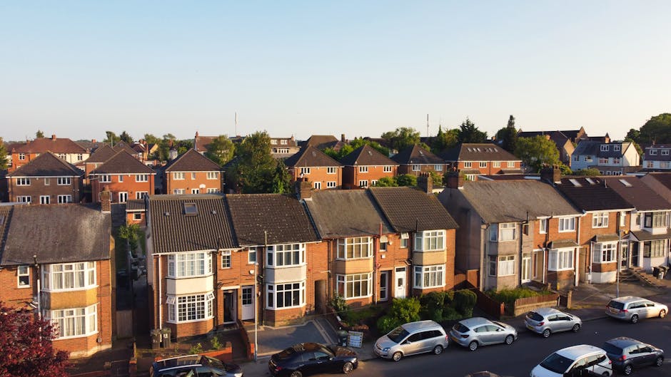 An aerial view of a suburban residential street with two rows of brick terraced and semi-detached houses, each with front gardens and driveways. Several cars, including sedans and hatchbacks, are parked along the street and on the pavements. The houses feature pitched roofs with tiles, some with chimneys, and a variety of window styles, including bay windows and sash windows. Visible items indicating packing and home relocation preparations include cardboard boxes and furniture blankets placed on the pavement near some houses, suggesting ongoing or planned furniture transport. The environment is well-lit with natural daylight, and the background shows a mix of detached homes and trees, indicating a typical UK neighbourhood. This scene aligns with professional house removals or property moving services, as provided by Man with Van Neasden, showcasing the logistics involved in arranging home relocation, loading furniture and boxes onto vehicles, and managing street access for efficient moving processes.
